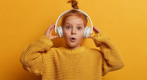 Surprised ginger kid listens audio track in headphones, impressed by loud sound, opens mouth with wonder, wears oversized knitted sweater, isolated on yellow wall. Children and hobby concept